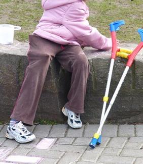 A photo of a person from the shoulders down. They are sitting on a rock bench and wearing purple pants and a pink coat.  Their colorful forearm crutches are resting on the stone bench next to them.
