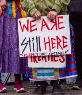 women with a sign that reads "we are still here - respect our existence"