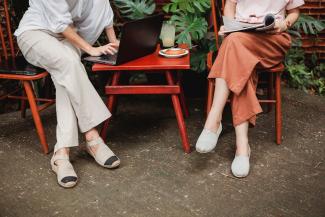 two people sitting outside at a red table and chairs while one types on a laptop